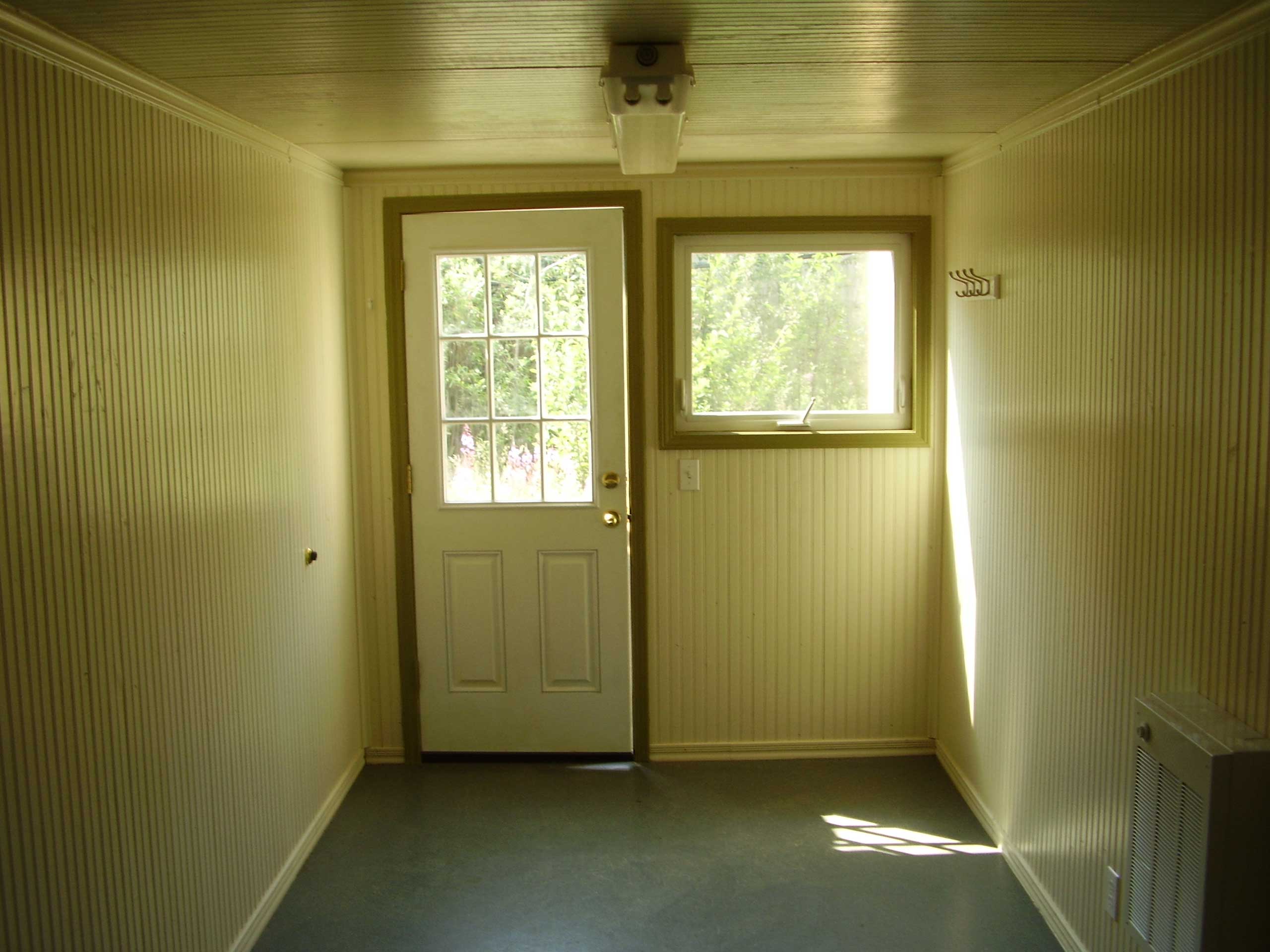 Office entry door, window-panes for natural lighting, painted interior wood-panelling with commercial carpet, wall heating system, and fluorescent lighting fixtures for a comfortable working environment for employees as either a temporary on-site office space, or off-site field office.